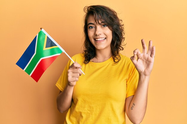 Young hispanic woman holding south african rand banknotes doing ok sign with fingers, smiling friendly gesturing excellent symbol