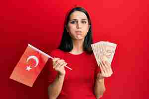 Free photo young hispanic woman holding turkey flag and liras banknotes puffing cheeks with funny face. mouth inflated with air, catching air.