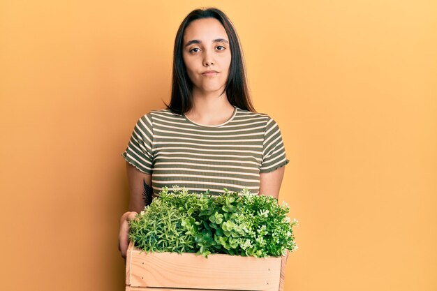 Young hispanic woman holding wooden plant pot relaxed with serious expression on face. simple and natural looking at the camera.