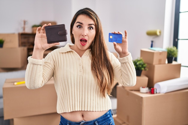 Young hispanic woman moving to a new home holding wallet and credit card afraid and shocked with surprise and amazed expression fear and excited face