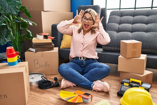 Free photo young hispanic woman moving to a new home sitting on the floor smiling cheerful playing peek a boo with hands showing face. surprised and exited
