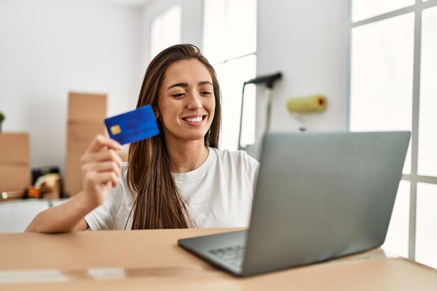 새 집에서 신용 카드와 노트북을 사용하여 자신감 있게 웃고 있는 젊은 히스패닉 여성