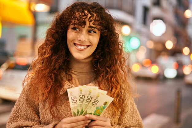 도시에서 이스라엘 세겔 지폐를 들고 행복하게 웃고 있는 젊은 히스패닉 여성
