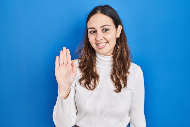 Young hispanic woman standing over blue background waiving saying hello happy and smiling friendly welcome gesture