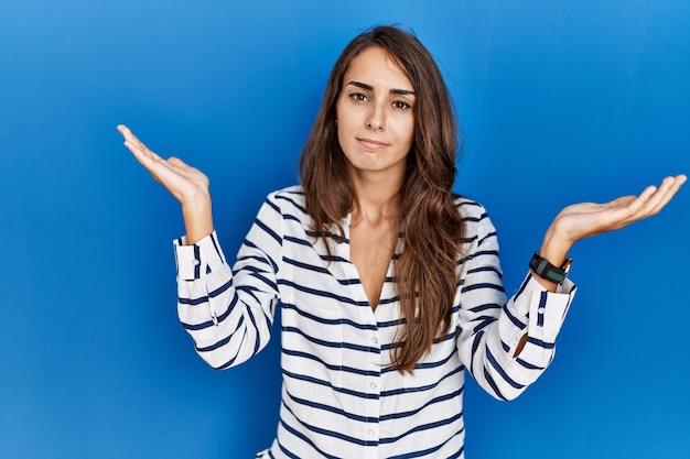 Free photo young hispanic woman standing over blue isolated background clueless and confused expression with arms and hands raised doubt concept