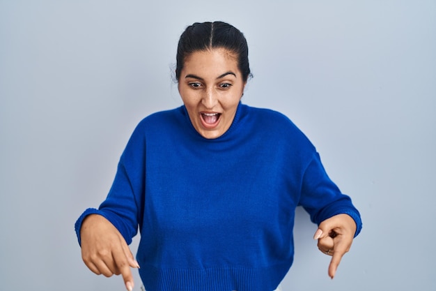 Young hispanic woman standing over isolated background pointing down with fingers showing advertisement, surprised face and open mouth