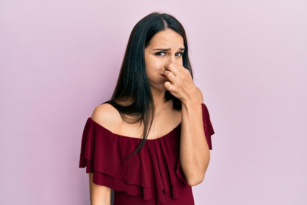 Young hispanic woman wearing casual clothes smelling something stinky and disgusting, intolerable smell, holding breath with fingers on nose. bad smell