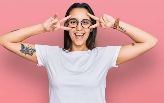 Free photo young hispanic woman wearing casual white t shirt doing peace symbol with fingers over face smiling cheerful showing victory