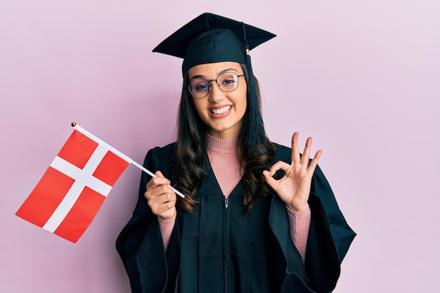 Foto gratuita giovane donna ispanica che indossa l'uniforme di graduazione che tiene la bandiera della danimarca facendo segno ok con le dita sorridenti gesti amichevoli simbolo eccellente