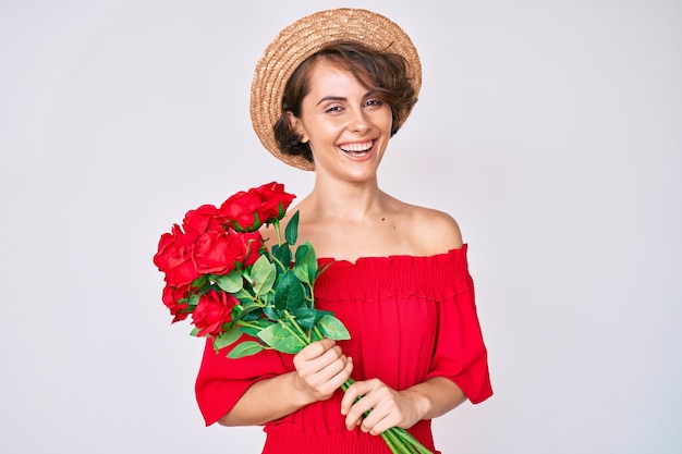 Free photo young hispanic woman wearing hat holding flowers looking positive and happy standing and smiling with a confident smile showing teeth