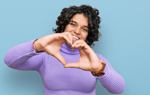 Young hispanic woman with curly hair wearing casual clothes smiling in love doing heart symbol shape with hands romantic concept