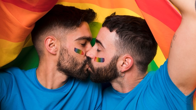 Молодая гомосексуальная пара мужчин целуется на флаге ЛГБТ