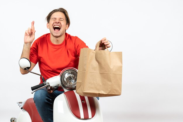 Ragazzo giovane corriere emotivo promettente in uniforme rossa che si siede sul motorino che dà il sacchetto di carta sul muro bianco