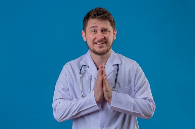 Young hopeful worried doctor wearing white coat and stethoscope holding hands in pray over isolated blue background