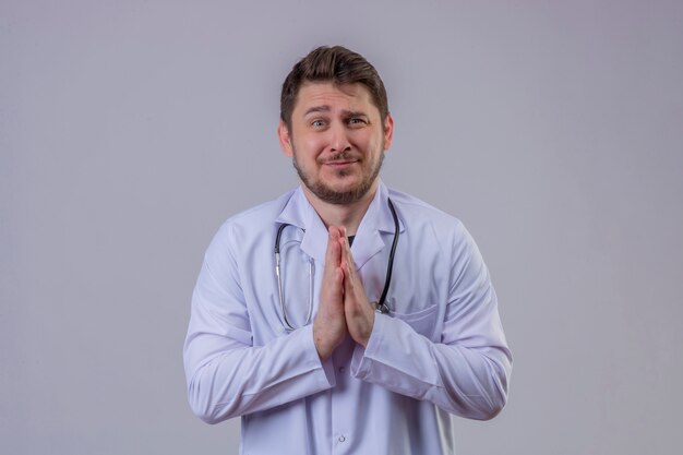 Young hopeful worried doctor wearing white coat and stethoscope holding hands in pray