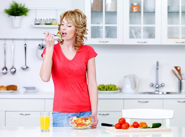 Молодая домохозяйка ест салат из овощей на кухне - в помещении
