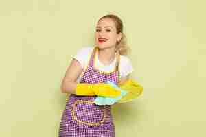 Бесплатное фото Молодая домохозяйка в рубашке и фиолетовой накидке с желтыми перчатками сушит тарелку на зеленом