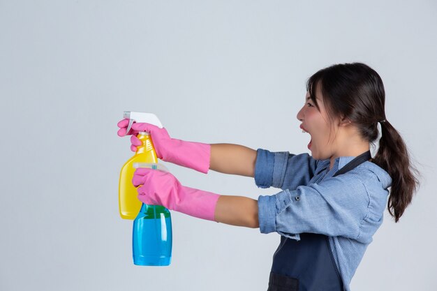 Молодая домохозяйка носит желтые перчатки во время уборки с продуктом чистой на белой стене.
