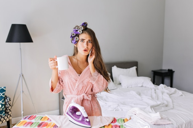 Young housewife in pink bathrobe at home at ironing clothes. She speaking on phone and holding a cup. She has curler on hair, looks astonished .