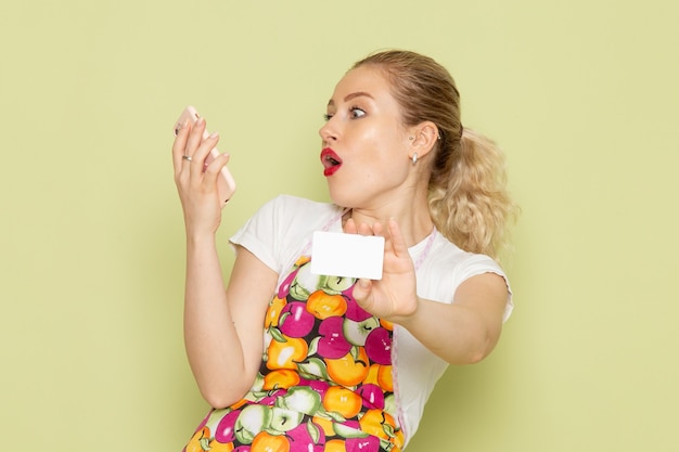 Free photo young housewife in shirt and colored cape holding white card and phone on green