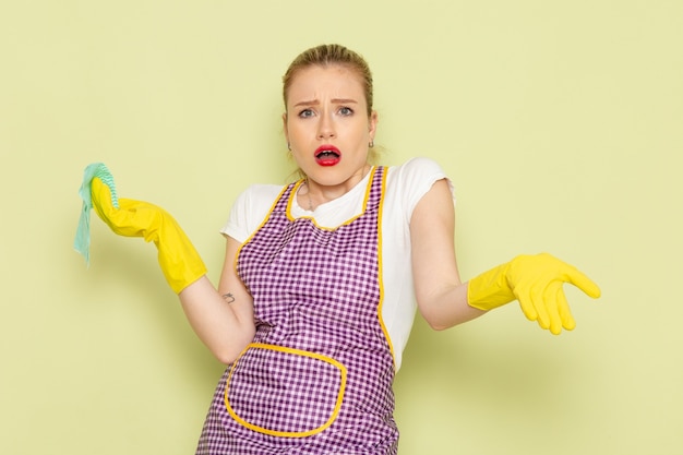 Free photo young housewife in shirt and purple cape with yellow gloves holding green rag confused on green