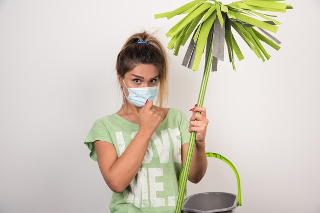 Young housewife with facemask holding mop and looking front on white wall. 