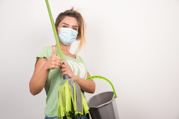 Young housewife with facemask holding mop making thumbs up.