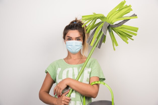 Young housewife with facemask holding mop on white wall. 