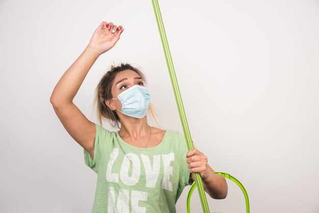 Young housewife with facemask holding mop on white wall. 