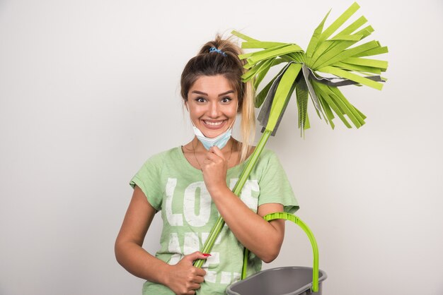 Young housewife with facemask holding mop with happy expression on white wall. 