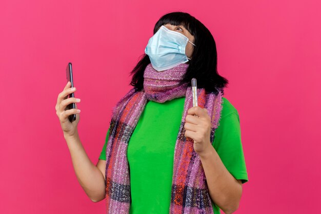 Young ill caucasian girl wearing mask and scarf holding thermometer and mobile phone looking up isolated on crimson background with copy space