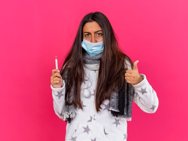 Young ill girl looking at camera wearing medical mask with scarf holding thermometer showing thumb up isolated on pink background