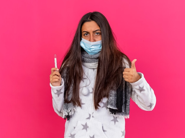 Young ill girl looking at camera wearing medical mask with scarf holding thermometer showing thumb up isolated on pink background