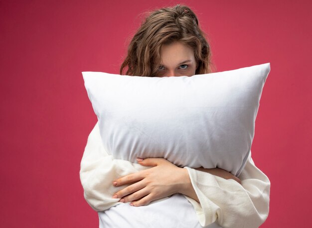 Young ill girl looking straight ahead wearing white robe hugged pillow isolated on pink