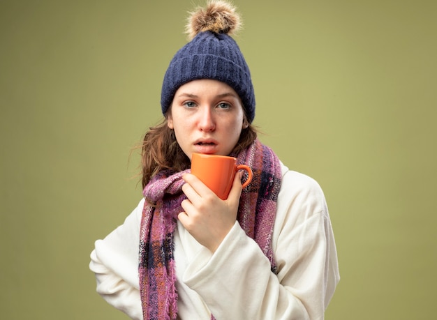 Young ill girl looking straight ahead wearing white robe and winter hat with scarf holding cup of tea putting hand on hip isolated on olive green