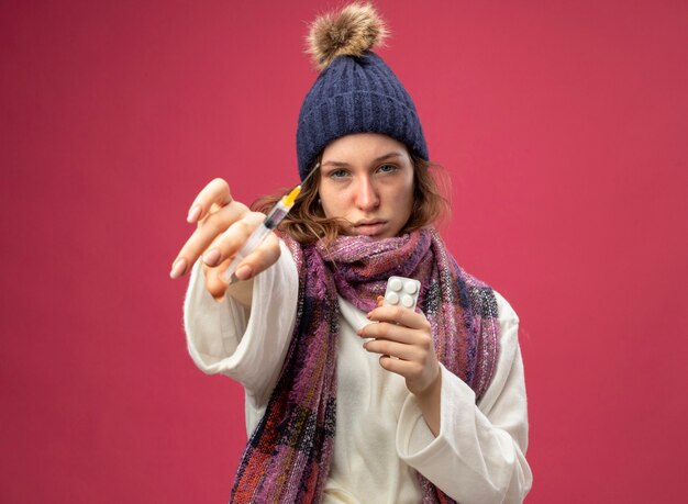 Молодая больная девушка смотрит прямо перед собой в белом халате и зимней шапке с шарфом, держит таблетки и держит шприц, изолированный на розовом