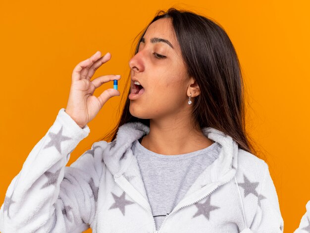 黄色の背景で隔離の口に錠剤を入れている若い病気の女の子
