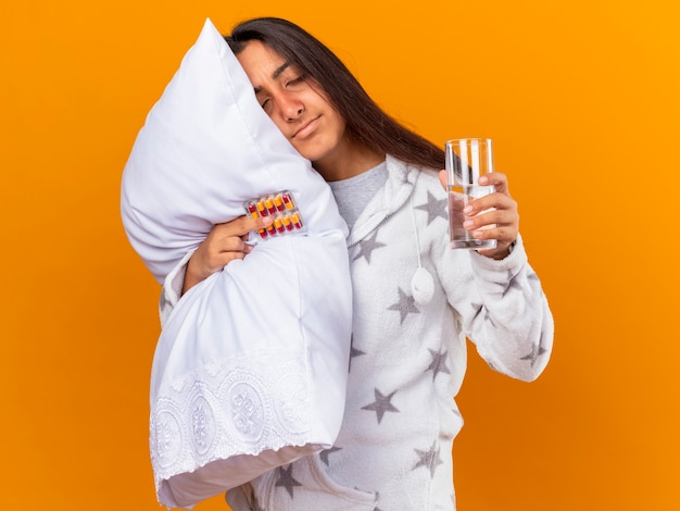 Young ill girl with closed eyes hugging pillow holding pills with glass of water isolated on yellow background