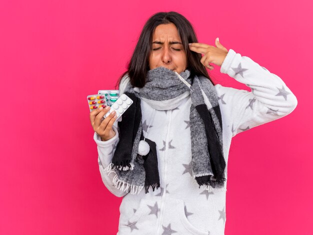 Young ill girl with closed eyes wearing and covered mouth with scarf holding pills putting thermometer in mouth showing suicide with pistol gesture isolated on pink