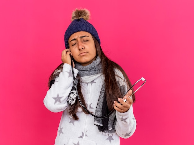 Foto gratuita giovane ragazza malata con gli occhi chiusi che indossa il cappello invernale con la sciarpa che tiene lo stetoscopio sull'orecchio isolato sul colore rosa