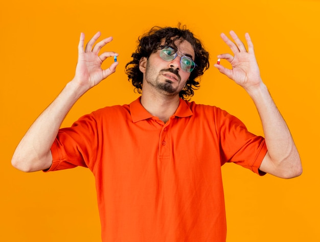 Free photo young ill man wearing glasses holding and looking at medical capsules isolated on orange wall