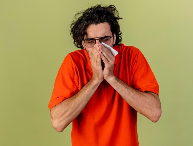 Young ill man wearing glasses holding napkin keeping hands on mouth and sneezing isolated on olive green wall with copy space