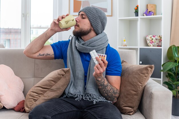 young ill man wearing scarf and winter hat sitting on sofa in living room holding packs of medical pills drinking cup of tea