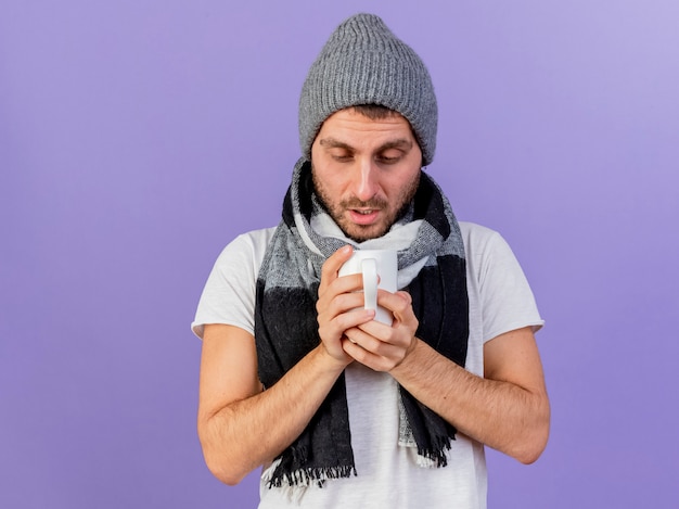 Young ill man wearing winter hat with scarf holding and looking at cup of tea isolated on purple background