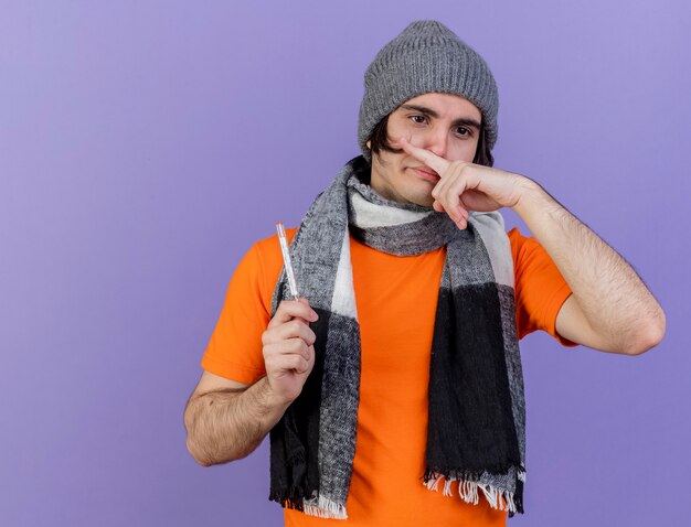 young ill man wearing winter hat with scarf holding thermometer wiping nose with finger isolated on purple