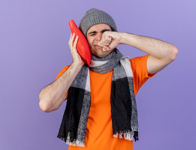 young ill man wearing winter hat with scarf putting hot water bag on cheek wiping face with hand isolated on purple