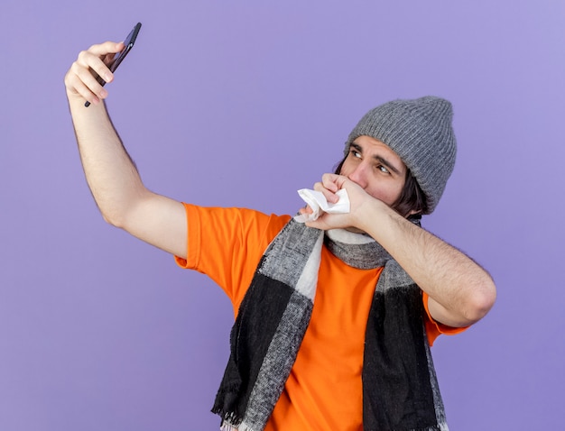 Бесплатное фото Молодой больной мужчина в зимней шапке с шарфом делает селфи и вытирает нос рукой, изолированной на фиолетовом фоне