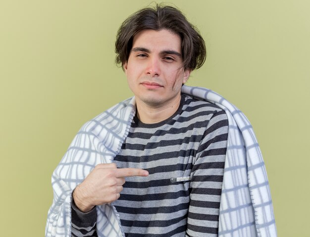 young ill man wrapped in plaid points at himself isolated on olive green