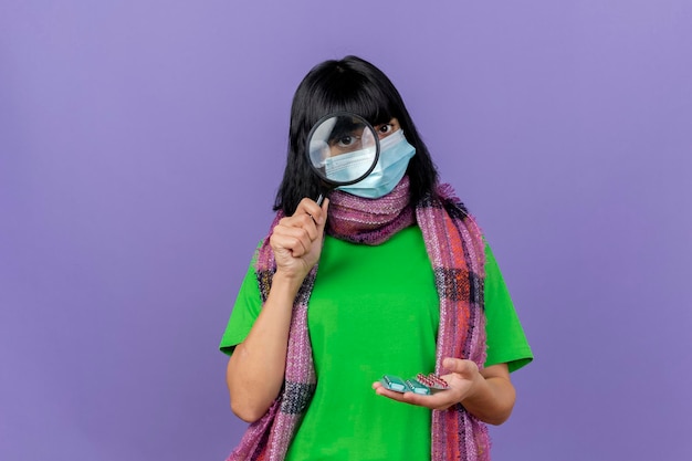 Young ill woman wearing mask and scarf holding packs of capsules looking at front through magnifying glass isolated on purple wall with copy space