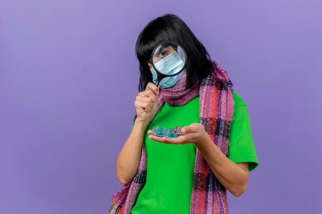 마스크와 스카프를 착용하는 젊은 아픈 여자 복사 공간이 보라색 벽에 고립 된 돋보기를 통해 전면을보고 캡슐의 팩을 들고